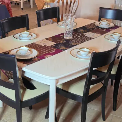 White rectangular dining table with six black wooden chairs featuring upholstered seats, set with a patterned table runner, crystal vase,...