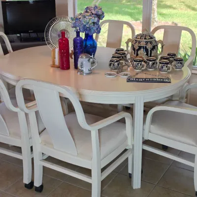 White painted oval dining table with six matching caster-wheeled armchairs, decorated with various glassware, a punch bowl set, and...