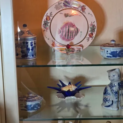 Collection of decorative porcelain and glass collectibles displayed in a white curio cabinet, featuring a hand-painted fish plate, blue...
