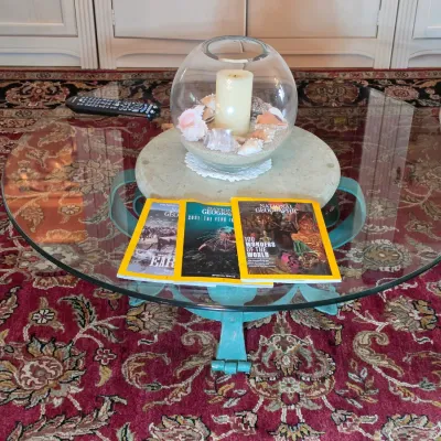 Round glass-top coffee table with a teal-painted metal base, displaying three National Geographic magazines and a glass candle holder...