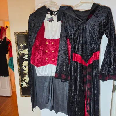 Two Halloween costumes hanging on a door, including a vampire outfit with a red vest and cape, and a black velvet witch or sorceress gown...