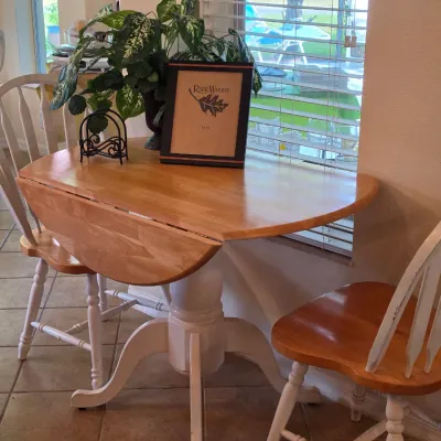 Country-style drop-leaf dining table with a natural wood top and white pedestal base, accompanied by two matching spindle-back chairs.
