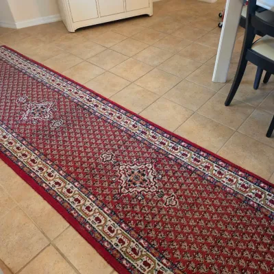 Traditional Persian-style runner rug featuring a red field with geometric medallions and a multi-layered border, shown on a tiled floor.