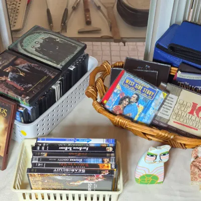 Assorted DVDs and music CDs in plastic bins and a wicker basket, featuring titles like "The Lord of the Rings," "Training...