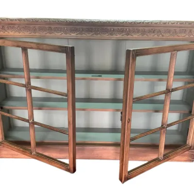 Vintage wooden display cabinet featuring glass-paned double doors, carved leaf detailing along the top frieze, and three interior shelves.