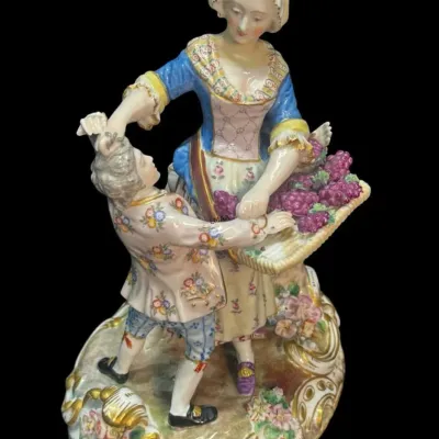 Antique German porcelain figurine depicting a man and woman with a basket of grapes, featuring hand-painted details and gold accents on a...
