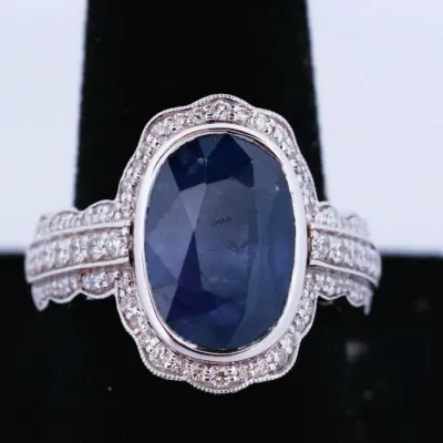 Vintage-style white gold ring featuring a large oval-cut blue sapphire center stone, surrounded by a scalloped halo of small round...