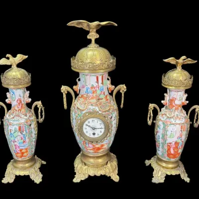 Three-piece French or Chinese export style garniture set featuring a central porcelain urn clock and two matching side urns, decorated...