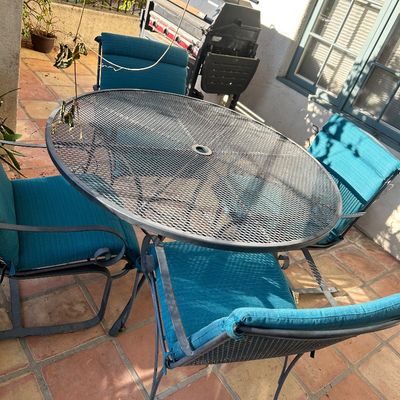 Wrought iron patio dining set featuring a round mesh-top table with an umbrella hole and four matching chairs with teal cushions.