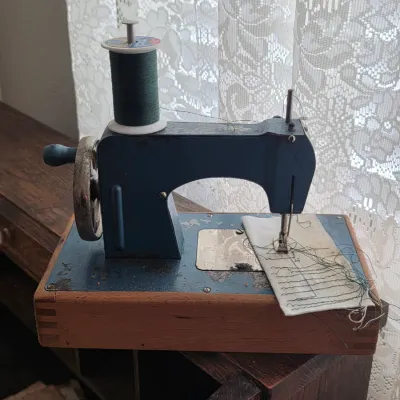 Vintage toy hand-crank sewing machine in blue metal, mounted on a wooden base with a spool of green thread.