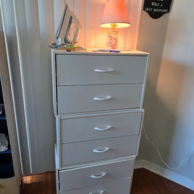 Stack of three white modular two-drawer storage units with curved plastic handles, topped with a small table lamp and a clothes iron.