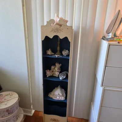 Vintage hand-painted wooden shelf unit with a distressed cream exterior, dark blue interior shelves, and decorative scrollwork,...