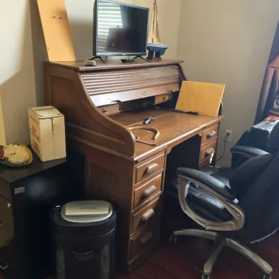 Vintage oak rolltop desk with multiple drawers, paired with a modern black leather office chair and various desk accessories.