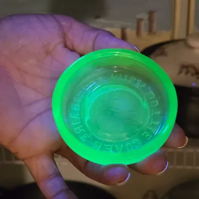 Vintage green uranium glass furniture caster cup glowing under a UV light, featuring embossed lettering along the rim.
