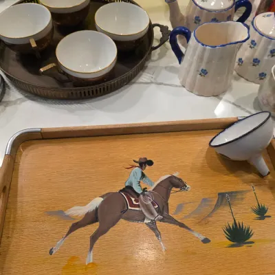Vintage mid-century wooden serving tray featuring a hand-painted cowboy on horseback, shown alongside assorted tea sets and a small...