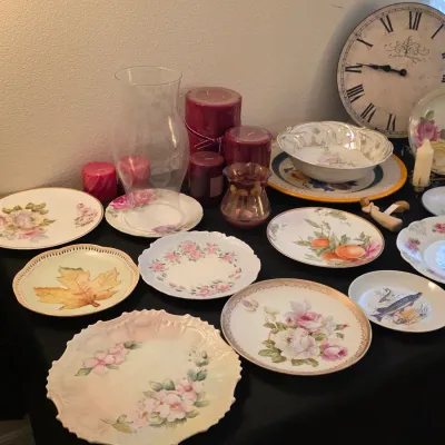 Assortment of vintage decorative plates with floral, leaf, and fruit motifs, displayed alongside a glass hurricane vase, red pillar...