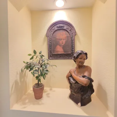 Decorative wall niche featuring a female bust sculpture, a framed portrait of a child in an ornate arch frame, and a small faux potted...