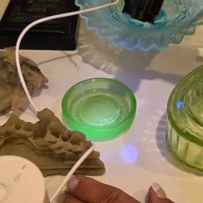 Small green uranium glass dish glowing under a UV flashlight, shown alongside other glassware and decorative items.