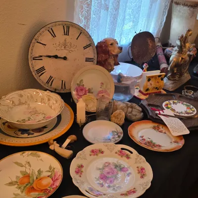 Assortment of vintage tabletop decor, including floral porcelain plates, a large Roman numeral wall clock, a ceramic dog figurine, and...