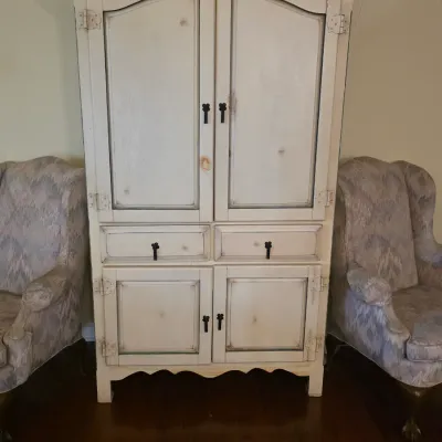 Distressed white wooden armoire with dark metal hardware, flanked by two upholstered wingback armchairs.