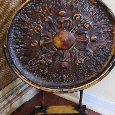 Decorative embossed metal plate on a matching metal stand, featuring a rustic bronze and gold finish with a central floral motif.