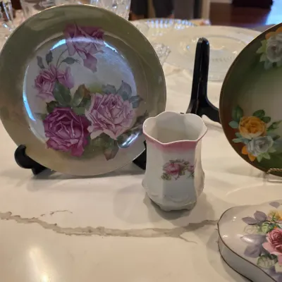 Vintage hand-painted porcelain plates and a small vase featuring delicate rose motifs, displayed on stands.
