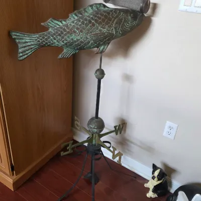 Vintage copper fish weathervane with a heavy green patina, mounted on a black metal tripod stand.
