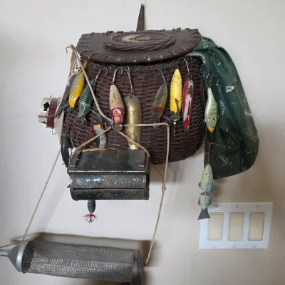 Vintage wicker fishing creel decorated with various antique lures, a metal bait box, and a green patterned bandana.