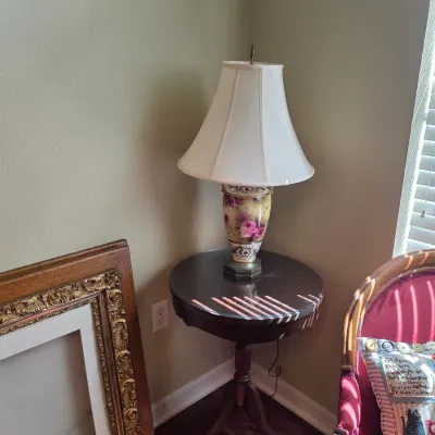 Vintage hand-painted porcelain table lamp with floral motifs and a white fabric shade, displayed on a dark wood tripod side table with a...