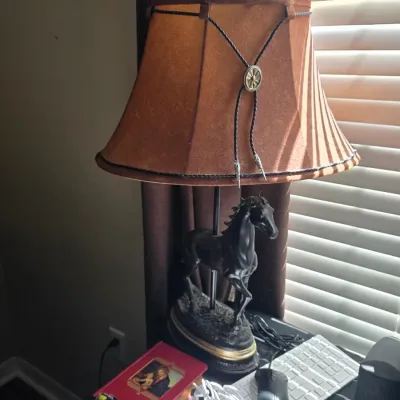 Western-themed table lamp featuring a black horse sculpture base and a faux-suede bell shade adorned with a bolo tie accent.