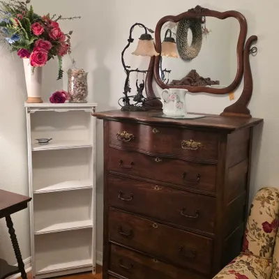Antique dark wood tallboy dresser with a matching swivel mirror, ornate brass hardware, and a serpentine top drawer, shown alongside a...