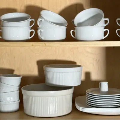 Collection of white ceramic tableware, including double-handled soup bowls, ribbed ramekins, soufflé dishes, and plates, arranged on...