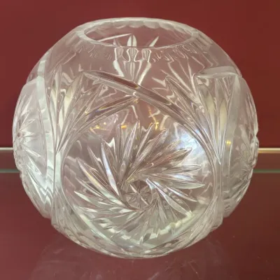 Vintage cut crystal rose bowl vase featuring a pinwheel and starburst pattern, displayed on a glass shelf against a red background.