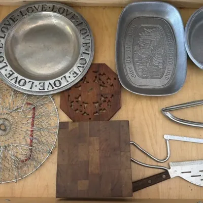 Assorted vintage kitchenware including pewter plates, a wooden trivet, a small butcher block cutting board, a collapsible wire basket,...