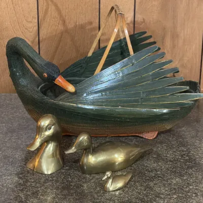 Sale Photo Thumbnail #197: Vintage woven wicker swan basket with painted green wings and orange beak, accompanied by three solid brass duck figurines of varying sizes.