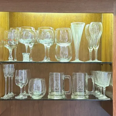 Assorted glassware collection displayed in a lighted wooden cabinet, including wine glasses, champagne flutes, beer mugs, and tumblers.