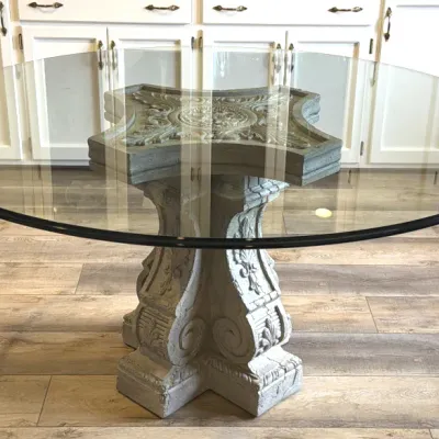 Round glass-top dining table featuring a thick, beveled edge supported by a classical-style cast stone pedestal base with ornate scrollwork.