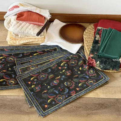 Assorted table linens including dark floral placemats, green and red cloth napkins, a woven charger plate, and various kitchen towels.