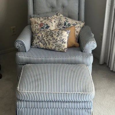 Sale Photo Thumbnail #106: Traditional wingback armchair and matching ottoman upholstered in blue and white ticking stripe fabric, styled with several floral accent pillows.
