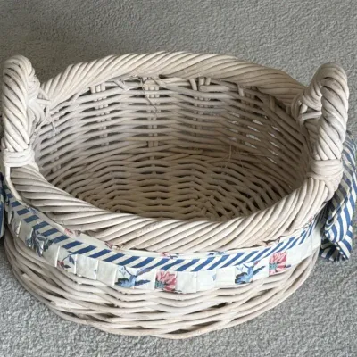 Sale Photo Thumbnail #101: Round whitewashed wicker basket featuring two woven handles and a decorative blue and white floral fabric trim with side bows.