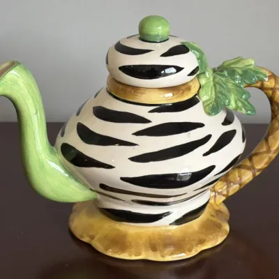 Whimsical ceramic teapot featuring a zebra-striped body, a green spout, and a palm tree-inspired handle with green leaves.