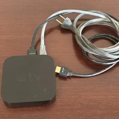 Apple TV media receiver console with power cord and HDMI cable, showing some light surface wear.
