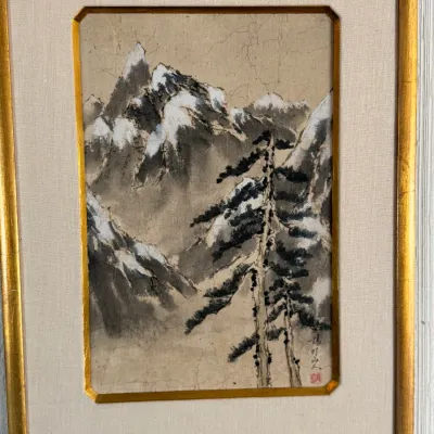 Framed East Asian ink and wash landscape painting depicting snow-dusted mountains and pine trees, presented in a gold-gilded frame with a...