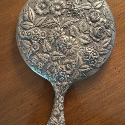 Vintage sterling hand mirror featuring an intricate, repoussé floral design with roses, daisies, and leaves covering the back and handle.