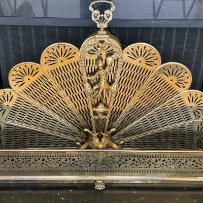 Vintage brass peacock-style folding fireplace screen with an ornate central figure and matching brass fender.