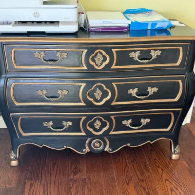 French Provincial style three-drawer bombe chest, painted black with gold-painted trim and ornate brass hardware.
