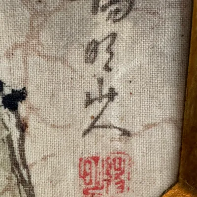 Close-up of an artist's signature and red seal stamp on a textured canvas or silk painting, framed in a gold-toned border.