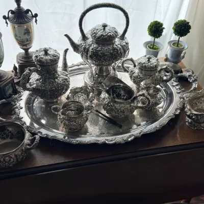 Ornate silver-plated tea and coffee service on a matching serving tray, featuring heavily embossed floral and scroll motifs, accompanied...