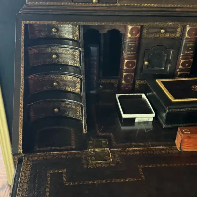 Antique secretary desk interior featuring ornate gold-painted borders, curved miniature drawers, and faux book spine compartments.