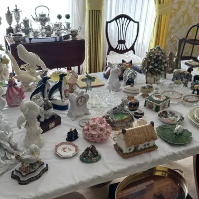 Large collection of decorative porcelain figurines, trinket boxes, and tableware displayed on a white tablecloth, featuring animal...